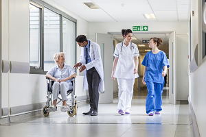 Two female Nurses are walking in a Hospital Hallway past a male Physician who is talking to an elderly female patient that is sitting down in a wheelchair